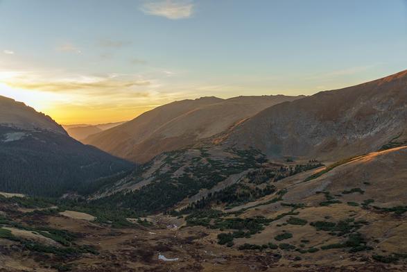 Landscape photo. The sun is just rising behind a mountain ridge at the left, viewed from the Trail Ridge Visitor Center in Rocky Mountain National Park. A glacier-carved bowl valley spreads out below the visitor center, looking east. October 2015.