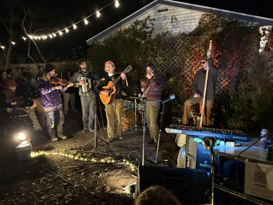 Six piece folk band, playing outdoors in the nighttime. Left to right: masc fiddle player in shiny purple jacket, cute masc accordion player with curly hair, me, center stage playing a jumbo 12-string, petite banjo player, tall man in sunglasses on washtub bass; smiling drummer behind all of us