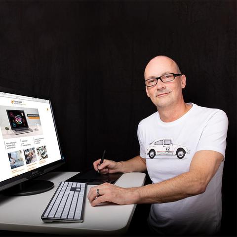 Stefan Lamb steht an einem höhenverstellbaren Schreibtisch. Er trägt ein weißes T-Shirt mit einem Auto, einem VW Käfer, drauf. Auf dem Schreibtisch befindet sich eine Tastatur, ein Grafiktablet und ein Monitor, auf dem die Website von Stefan Lamb zu sehen ist. Stefans linke hand liegt vor der Tastatur, mit der rechten hält er einen Stift fest.