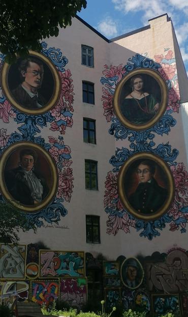 An einer Hauswand sind 4 grosse Portraits gemalt in ovalen Rahmen mit Blumenverzierung darum.  Zwischen den Bildern ist eine Reihe schmaler kleiner Fenster.