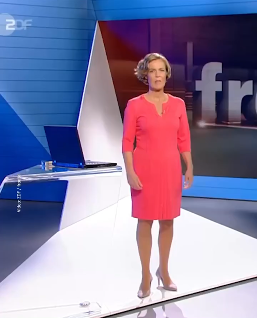 A woman wearing a pink dress and high heels stands in a television studio, at a desk, picks up a hammer, and smashes a laptop. She then picks up a laptop, drops it onto the studio floor, and stomps on it with her shoe.