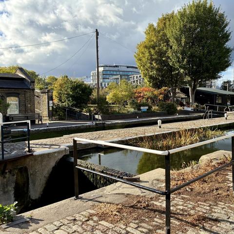 A lock on the grand union canal with cobblestones in foreground and brick buildings and greenery (and a red flowering bush) behind it, cloudy skies in background.