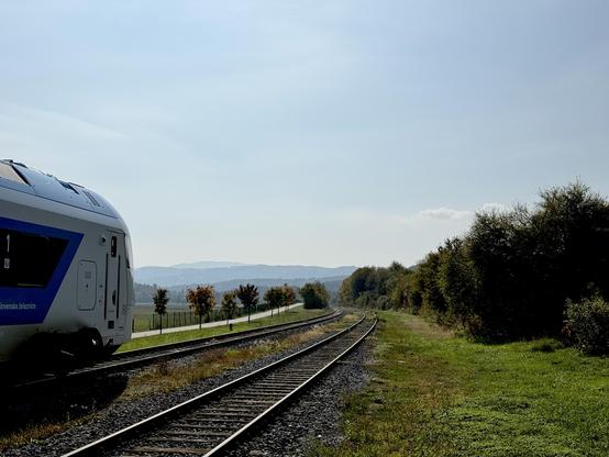Two tracks lead off into the distance in a gentle curve to the left. A white and blue train is peeking into the picture on the left edge. Rolling hills are in the far away distance.