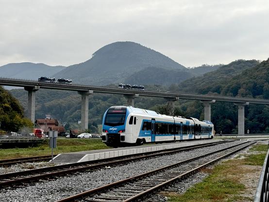 A white and blue train – a Slovenian diesel Flirt – is waiting at a forsaken platform. In the back a huge motorway bridge is crossing the picture with two lorries and some cars on it. Down on the valley floor is a normal road that is also very busy.
