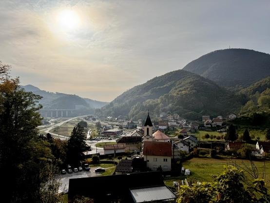 View over a village in a valley from above. Straight ahead is a church, beyond are maybe two, three dozen houses. Far back and really small is a railway station with a train waiting. Beyond are round mountains.