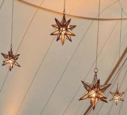 A color photo of four differently sized Moravian stars hanging on chains from a tent ceiling. The sides of the tent have a warm glow to the white tent.