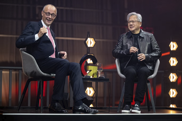 Foto. Kanzler Merz und Jensen Huang beim "Investitionsgipfel" auf der Bühne. Zwischen ihnen auf einem Tischchen die Weingläser, mit denen sie sich zugeprostet haben, und ein großes G, dessen Bogen schwarz und rot designt ist, der untere Haken aber in Form eines goldenen Pfeiles, der - natürlich - nach rechts oben weist.
