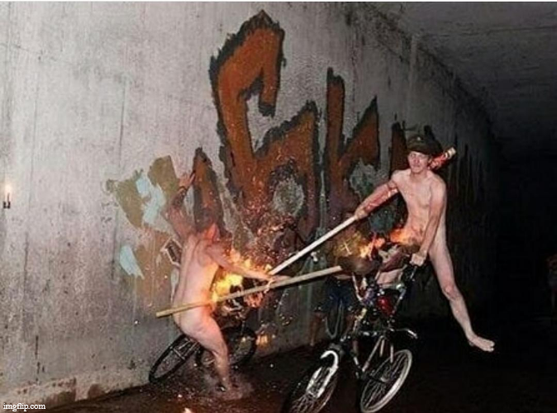 Two naked people jousting on bicycles, with lances that are on fire (maybe mops with the mop heads burning?).  They are  in some kind of concrete culvert or underpass.  There is dingy water on the ground, and graffiti on the wall behind them.  One is on a tall bike, the other a conventional bike.  They have just made contact, and it looks like both of them are probably going to fall.