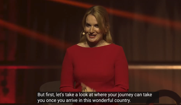 Screenshot. Bissig guckende Moderatorin in rot vor dem roten Hintergrund (schwarz und rot waren die Leitfarben des Events anscheinend - wegen Groko und weil das Gold in Form von Investitionen ja noch kommen soll). Caption: "But first, let's take a look at where your journey can take you once you arrive in this wonderful country."