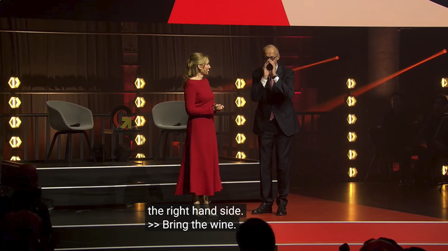Screenshot Event, Moderatorin und Merz auf der Bühne, Huang wird gerade angekündigt, Merz formt Hände zu einem Trichter vor dem Mund und ruft ihm etwas zu. Caption: "...the right hand side. >> Bring the wine!"