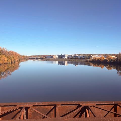 A calm river reflects the clear blue sky, autumn-colored trees, and distant buildings.