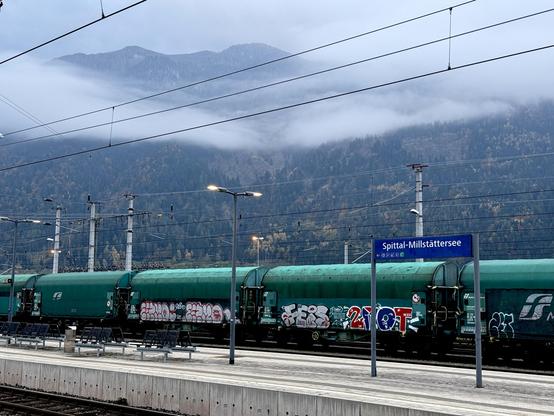 View across a railway platform with the sign Spittal-Milstättersee towards mountains with clouds halfway up.