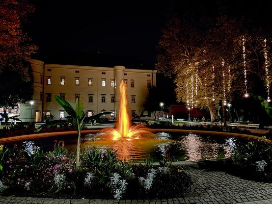 A round pond at night with a fountain lit in orange light. Beyond is a large old white building. There are also tree with light chains hanging from them.