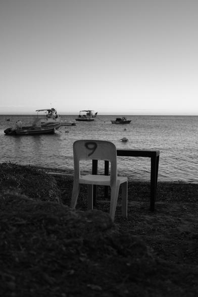 A black an wide picture of a single seat and a table facing the sea. The picture was taken at a beach which is visible in the foreground, while 4 smaller boats can be seen in the background.