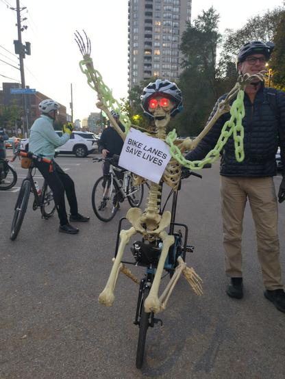 A skeleton with glowing red eyes and glow in the dark chains mounted on the front of a bike with a sign saying BIKE LANES SAVE LIVES