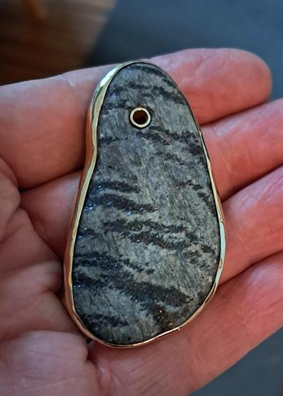Resting on a hand is a small flat stripey piece of schist. The stone has been bounded with metal; a hole drilled at one end has been lined with metal. Small crystals in the rock reflect light.