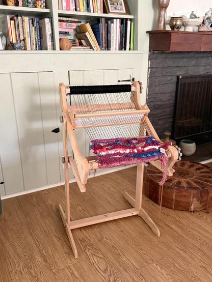 A 16” rigid heddle loom on a stand in front of my bookshelf. The loom has colorful yarn partially woven into a textile piece (by the previous loom owner.) You can see part of a fireplace mantle off to the side in the background. There’s also a round leather ottoman in the ground behind the loom.