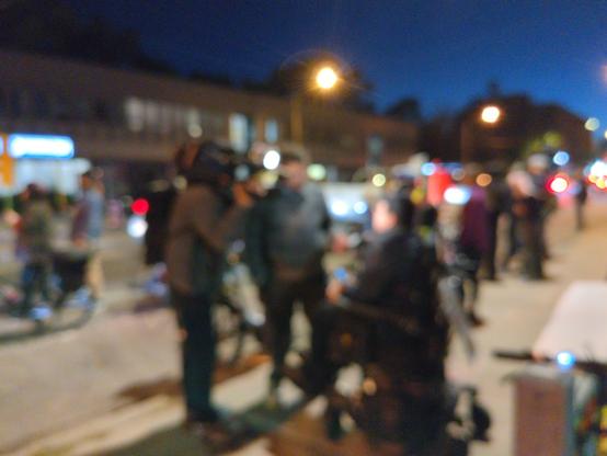 A camera operator and a reporter interview a protester who is in a motorized wheelchair. Photo blurred for privacy.