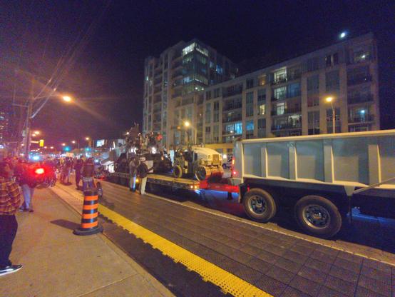 A large dump truck towing other machinery, with protesters around it.