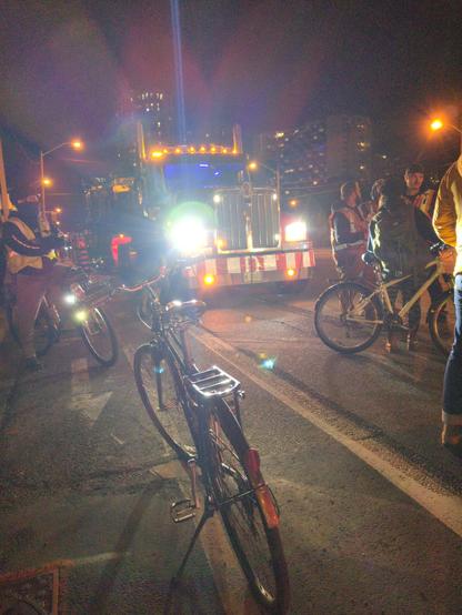 A few cyclists in front of the bright lights of the second truck, and also construction workers in safety vests.