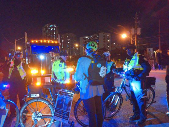Cops sternly telling protester (backpack, fluorescent helmet) to step off, protester staying put. Truck (empty) in background
