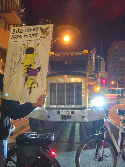 A protester holds a hand drawn sign saying "Bike lanes save moms" with a small Pikachu riding behind a large Pikachu. There is another Pokemon in the upper right hand corner (Eevee?) In the background is a dump truck being blocked in.