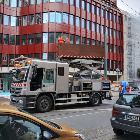 Quadratisches Foto. Vom Bürgersteig aus blickt die Kamera auf ein silberfarbenes Fahrzeug mit einer Hebebühne hintendrauf. Auf der Hebebühne ist ein hölzerner Kasten, in dem zwei Menschen mit orangenen Warnwesten stehen. Die Warnleuchten vorn oben des Fahrzeugs sind an. Hinter dem Fahrzeug stehen noch zwei Menschen mit den gleichen Warnwesten. Auf der anderen Straßenseite ist ein modernes, dunkelrotes Bürogebäude.