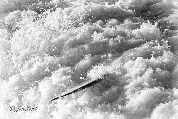 This is a black and white water sports photo in landscape format of the rear of a surfboard protruding out of a brilliant white and swirling mass of foaming surf. Hermosa Beach (2014).
The entire image is overflowing with the maelstrom of boiling surf from a breaking Pacific wave. The foaming water is brightly lit by the still low morning sun. Below the centre of the photo is the surfboard. The camera is located a few metres above and a few metres away from the board. Only the back and part of the mid section is visible, the remainder is hidden beneath the angry surf. The board is angled downwards towards the lower left corner, with it's edge angled towards the camera so little of the top or bottom surfaces are in view other than the one or two of the shark like fins on the underside of the board.