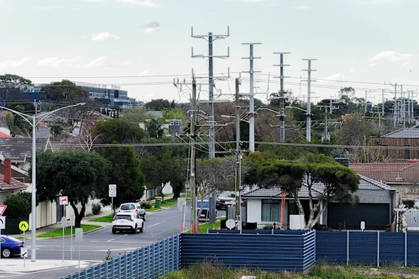 Suburban street with cars and single‑story houses dominated by tall electrical transmission poles and multiple overhead power lines behind a blue corrugated fence.