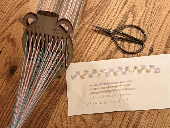 A pottery-shaped mini rigid heddle with several strands of yarn in turquoise, pink, and gray. Nearby, a pair of scissors and a handwritten note detailing a color coding system for the yarn colors being used in the project. All of the items lie on a wooden table.