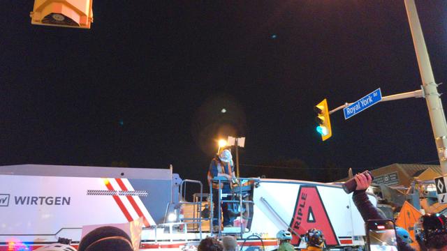A helmeted worker stands atop a Wirtgen cold milling machine at Royal York and Bloor. In the lower right you can see someone's arm holding up a cylindrical Bluetooth speaker.