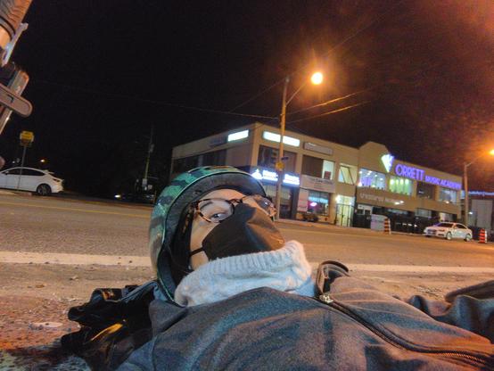 Me lying in the road, wearing a watermelon helmet, a black face mask, fleecy neck thing and jacket. There is a plaza on the other side of the road.