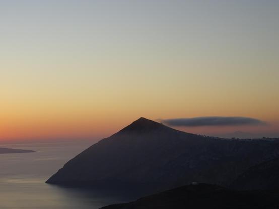 lever de soleil sur la pointe de l'île de Folégandros ans les Cyclades. On voit le ciel rose orangé , la mer douce et légèrement brumeuse et une montagne triangulaire qui émerge de l'eau, sombre