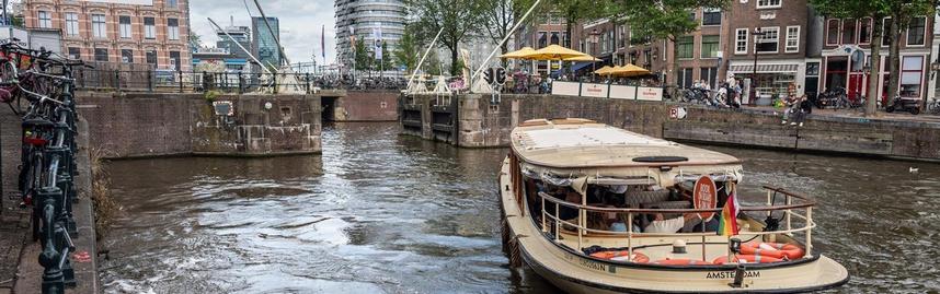 Een rondvaartboot vaart bij een stenen sluis. Langs het water staan fietsen en mensen, met gebouwen op de achtergrond.