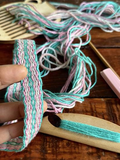 A close-up of my hand holding a woven band featuring pink and turquoise threads, with strands of yarn scattered in the background. A small wooden shuttle wrapped with turquoise yarn is also in view and you can see the small wooden heddle in the background. Everything lies on a wooden coffee table.