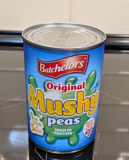 A can of Batchelors Original Mushy Peas with a blue label showing a green splash behind yellow and white lettering, plus badges reading "1 of your 5 a day," "High in Protein," and "British Grown and Packed" with a Union Jack heart. The can stands on a black glass cooktop.

(Description assisted by Claude AI.)