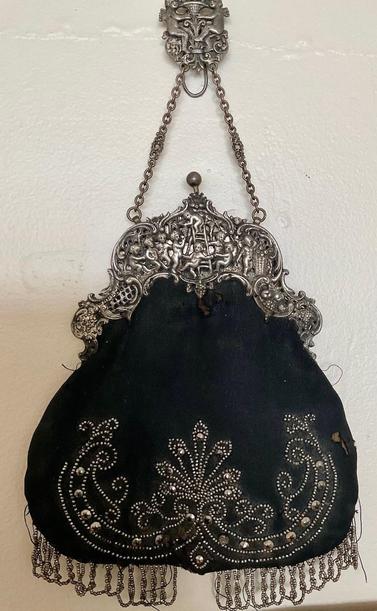 A black fabric chatelaine style with very tiny silvery crystal beads in a lineart style floral pattern and some larger studded crystal sequins. It almost looks western.  There’s some looping silvery crystal fringe at the bottom. The closure is large and curved and is carved in great detail like a frieze in sterling silver with children gathering fruits from trees. It has a silver chain that connects it to a chatelain loop decorated with a silver carved heraldic design of two putti and a cornucopia. The fabric and beading show wear, holes and missing beads.