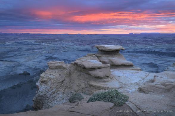 A color landscape photo of an almost surreal landscape at dawn. In the foreground are rounded sandstone formations at the edge to a deep drop into a vast basin of barren badlands with rounded mudd hills and long ridges. They sky above is cloudy and pre-sunrise light casts a glowing red color into the clouds.