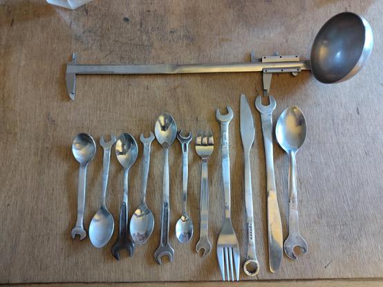 Sur une table en bois, des couverts (petites et grandes cuillères, fourchette, couteaux) qui ont chacun au bout de leur poignée un bout de clé plate, de différentes tailles et forme 
Au dessus, une louche dont le manche se termine par un pied à coulisse, dont la glissière est carrément sur la partie manche de la louche