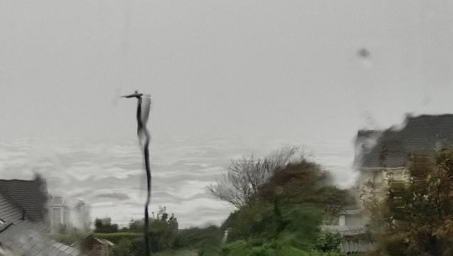 Looking through a rain covered window with lots of zoom, which makes everything a bit distorted. It suits the day we're having.

There's bits of houses visible, and lots of trees and hedges, and in the distance there's the sea, with big white topped waves on grey water. The sky is uniformly grey, and there appears to be nothing but grey out there.