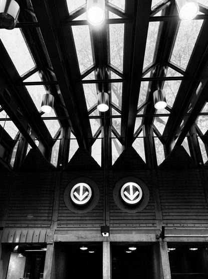 Black and white photograph taken inside a subway station and looking up, showing the rectangular glass windows that form the accordion-shaped ceiling, with cylindrical lights suspended from a metal structure and sunlight entering. Below, two round panels with arrows pointing downwards are hung on the concrete wall, textured with horizontal lines. Further down, the underground passages leading to the train, in the half-light, with round lights on the ceiling.

Photographie noir et blanc prise à l'intérieur d'une station de métro et regardant vers le haut, montrant les fenêtres vitrées et rectangulaires qui forment le plafond en forme d'accordéon, avec des lumières en forme de cylindres suspendues à une structure métallique et la lumière du soleil qui pénètre. En-dessous, deux panneaux ronds avec des flèches pointant vers le bas sont accrochées au mur de béton, texturé de lignes horizontales. Plus bas, les passages souterrains menant au train, dans la pénombre, avec des lumières rondes au plafond.