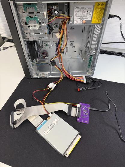 An open computer case showing internal components, including a power supply and various cables. A connected Floppy drive is positioned with a flat ribbon cable leading to a small purple circuit board, the greaseweazle.