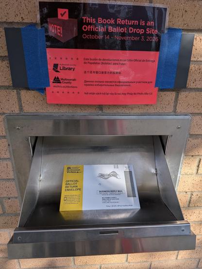 A red-and-black sign taped with blue painter's tape above a library book return reads "This Book Return is an Official Ballot Drop Site, October 14 - November 3, 2026," with the Multnomah County Library and Multnomah County logos. The stainless steel return chute hangs open in the brick wall, holding a yellow-and-white Multnomah Votes official ballot return envelope.

(Description assisted by Claude AI.)