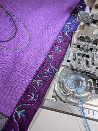 The edge of a quilt. It is bound in dark purple fabric, and a multicolored thread has made meandering stitches with bird feet shapes. The stitch pattern starts and stops nearly exactly perfectly.