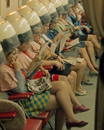 Looks like a seventies photo: about ten women are sitting next to each other at a beauty salon or hairdresser. They read magazines and have hairdryers on their hairdo's. All wear skirts.