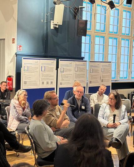 Teilnehmende des Dialogs für Cybersicherheit in einer Fishbowl-Diskussion. Ein Mann spricht in ein Mikrofon, Teilnehmende hören interessiert zu.