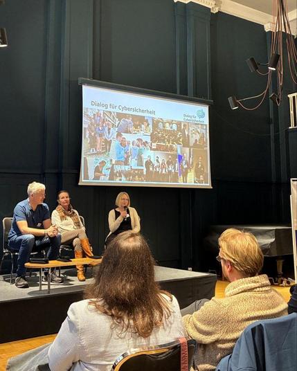 Teilnehmende einer Panel-Diskussion sitzen auf einer Bühne. Im Hintergrund ist eine Leinwand zu sehen, auf der eine Foto-Collage und der Schriftzug "Dialog für Cybersicherheit" abgebildet ist. Vorne sieht man ein paar Leute aus dem Publikum.