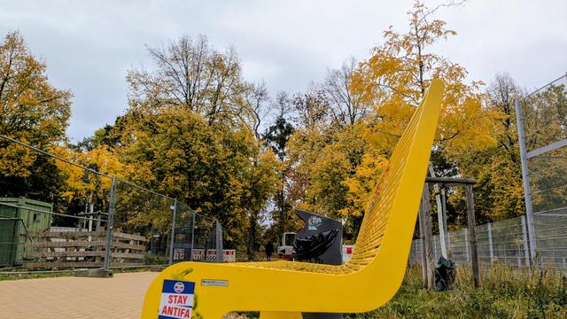 Knallgelbe Sitzbank im Park. Im Hintergrund tragen Bäume goldenes Herbstlaub. Berlin-typisch stehen hässliche Absperrgitter herum. Wenn's schon keine Mauer mehr gibt, wa?