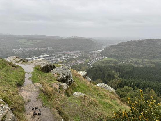 A grassy hill with rocks and a muddy path overlooking a town and a forested area under a cloudy sky. Taken from the Garth hill in Cardiff, Wales.