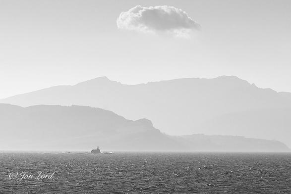 This is a black and white photo in landscape format of a modern submarine running on the surface with rising terrain in the background. Firth of Clyde (2017).
The dark waters of the sea rise from the base of the photo upwards to a little less than the quarter up level. On a poorly defined horizon and below and to the left of centre is our submarine, underway from left to right and just above the surface. The camera is located a few metres above the water with the submarine being around one km away. The all black vessel is viewed in profile with the starboard (right) side in view. Water can be seen washing over the casing (top of the hull) with a short sensor above the deck aft of the bow. Further aft is the large fin (sometimes called the sail or conning tower) that's located a little forward of midships with two or three raised signal masts, radars or periscopes. Further aft is more dark hull that is faired downwards towards the stern with a short stretch of water before the upper rudder is reached extending above the surface. A trail of white wake stretches behind at a similar length to the submarine. Beyond the vessel are two lines of undulating terrain, partially obscured by a summer's haze.
The closer ridge of terrain extending from the left edge of the photo and sloping downwards by the centre of the image. The further terrain is higher and stretches across the distant background with a near clear and cloudless sky above other than a single cumulus (cotton wool ball) cloud at the top of the photo above the centre.
